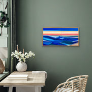 Artist Shilo Ratner painting Rise 20in x 40in, Modern water abstract painting of sunset with blue and red hues on a green wall, Modern interior with a colorful abstract painting on a gray wall, a table with a vase and books, and a chair.